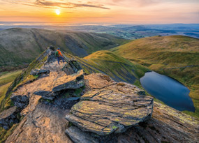 October - A hiker stands in an orange jacket and blue trousers on the peak of a mountain with jagged rocks. From left to right, the view includes rolling green hills, a golden sky at sunrise that beams down on to a large undisturbed lake.